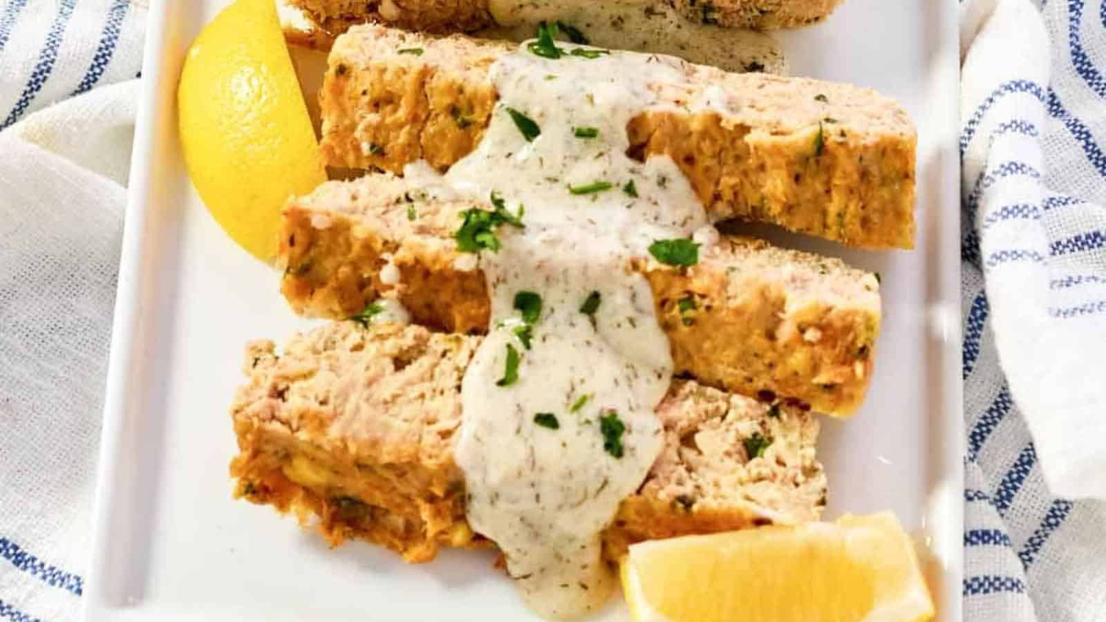 Slices of salmon loaf topped with creamy sauce and parsley, served with lemon wedges on a white plate.