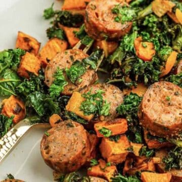 Sliced sausage, kale, and roasted sweet potatoes on a white plate with a gold fork.