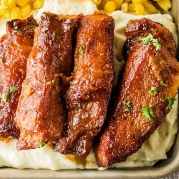 Glazed pork ribs on creamy mashed potatoes with corn in a square dish, garnished with parsley.