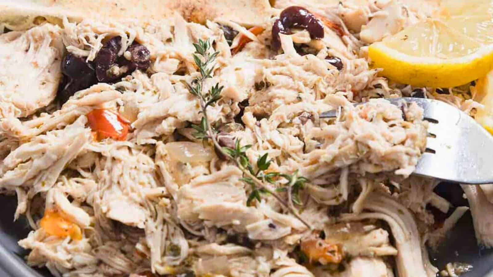 A plate of shredded chicken is garnished with herbs, black olives, and sliced cherry tomatoes. Two lemon wedges and pieces of flatbread accompany the dish. A fork is resting on the edge of the plate.