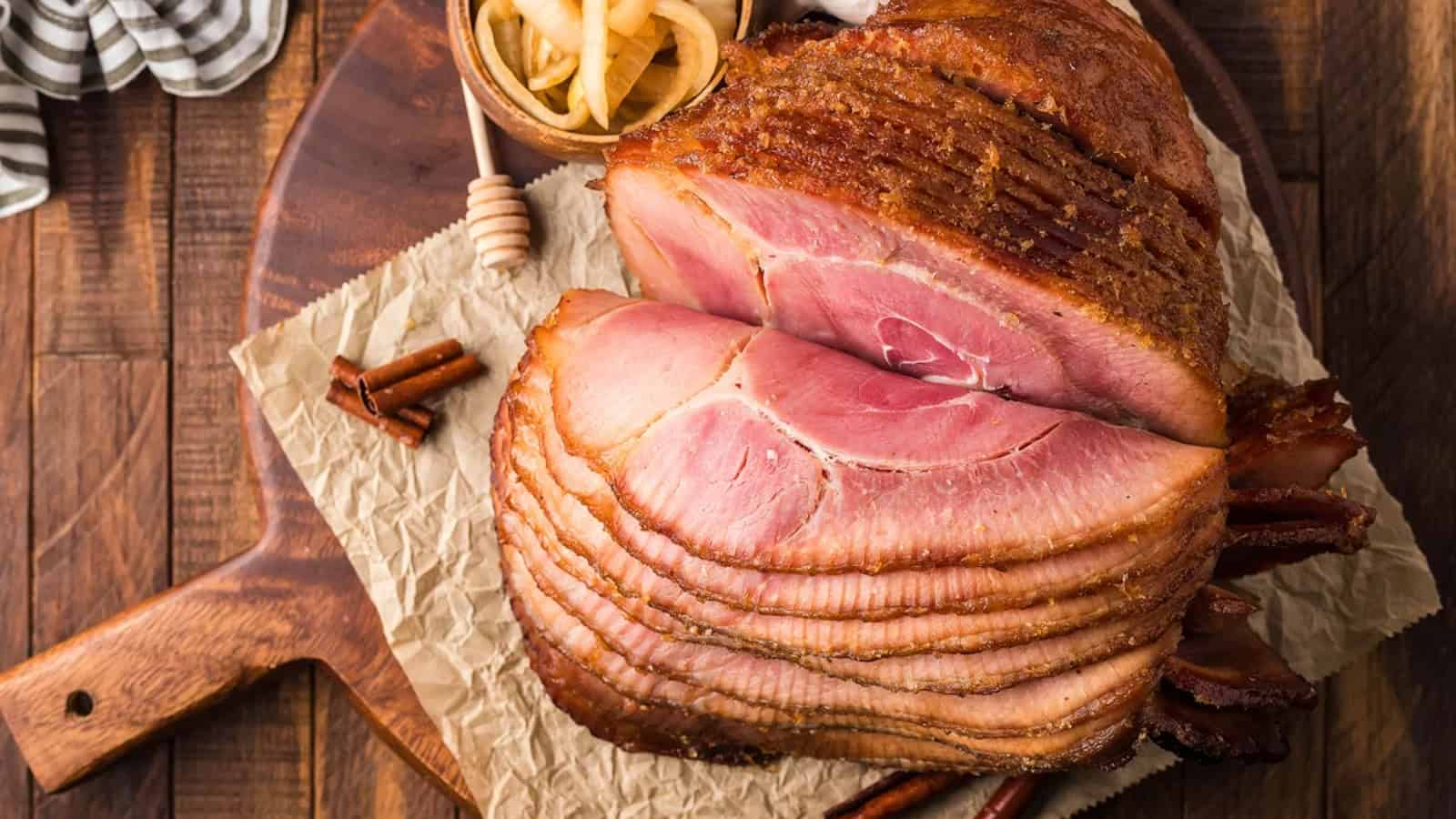 Sliced glazed ham on parchment paper with cinnamon sticks and a small bowl of pickled onions on a wooden board.
