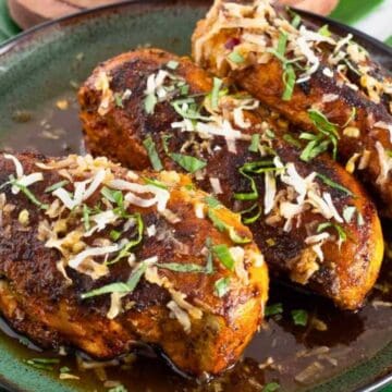 Three seasoned chicken breasts topped with shredded cheese and herbs on a dark plate with green garnishes.