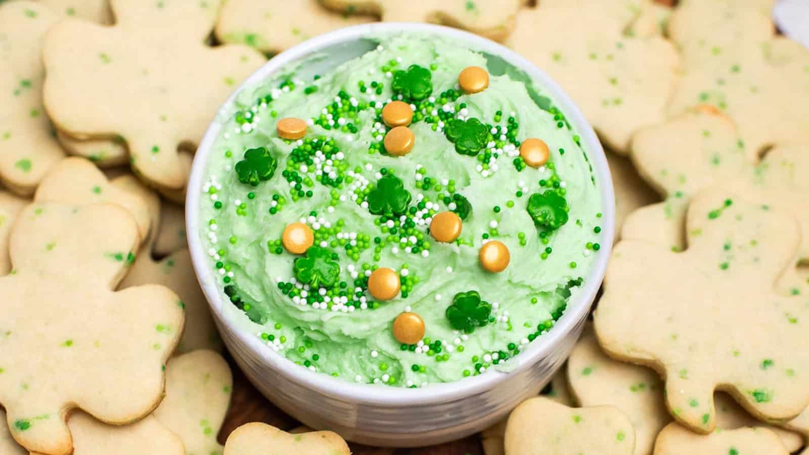 A bowl of green frosting with sprinkles, surrounded by shamrock-shaped cookies, makes a festive St. Patrick’s Day treat.