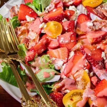 Salad with strawberries, cherry tomatoes, greens, and dressing on a white plate with gold utensils.
