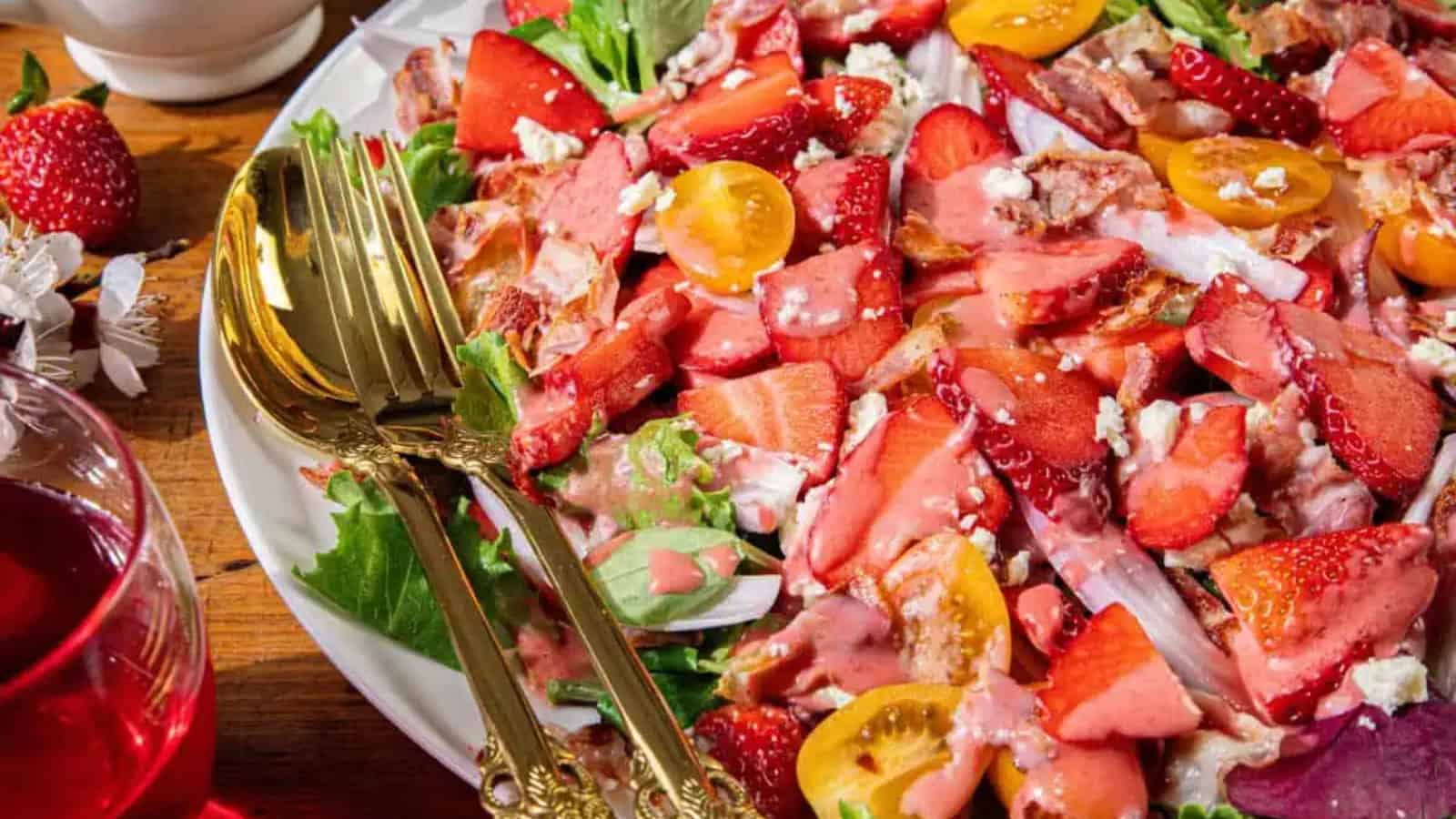 A vibrant spring salad with mixed greens, sliced strawberries, yellow cherry tomatoes, crumbled feta cheese, and bacon pieces, served with a pink strawberry vinaigrette. Gold utensils and a glass of red drink are beside the plate.
