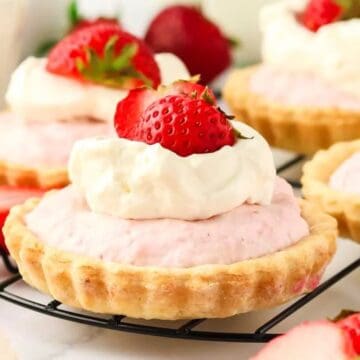 Mini strawberry tarts topped with whipped cream and fresh strawberries on a cooling rack.