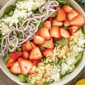 A salad bowl with spinach, sliced strawberries, red onions, feta cheese, and sliced almonds on a wooden table.