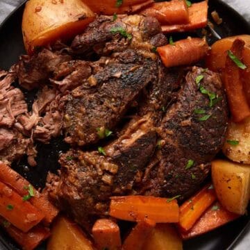 Plate with pot roast, potatoes, and carrots, garnished with herbs on a dark plate with a light cloth underneath—perfect for fans of comforting slow cooker recipes.