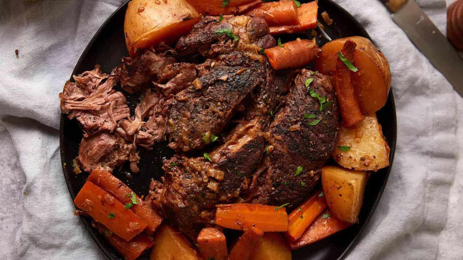 Plate with pot roast, potatoes, and carrots, garnished with herbs on a dark plate with a light cloth underneath—perfect for fans of comforting slow cooker recipes.