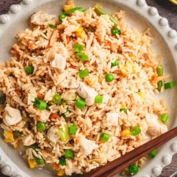 A plate of fried rice with vegetables, chicken pieces, and chopped green onions, with chopsticks on the side.