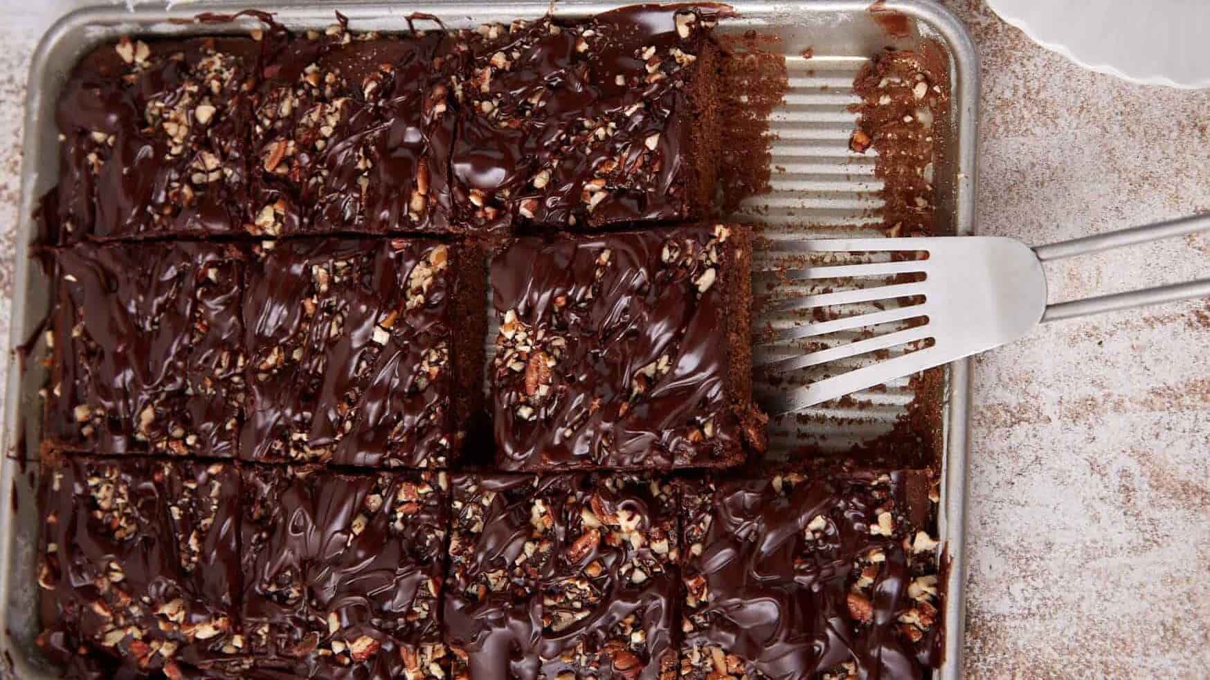 Chocolate sheet cake topped with nuts, cut into squares, with a spatula lifting one piece from the pan—a timeless treat inspired by vintage recipes.