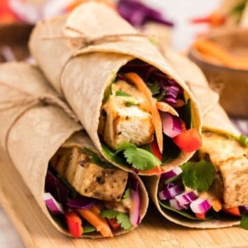 Three vegetable wraps with tofu, colorful veggies, and herbs, tied with twine, on a wooden board.