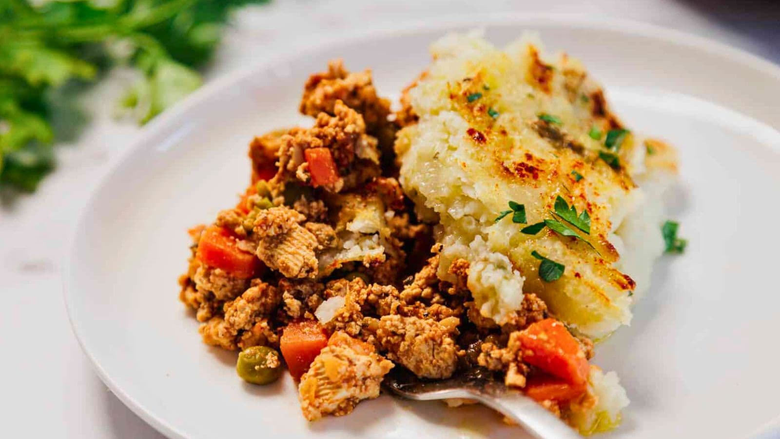 A serving of shepherd's pie with ground meat, peas, carrots, and mashed potatoes on a white plate.