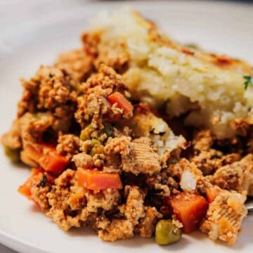 A serving of shepherd's pie with ground meat, peas, carrots, and mashed potatoes on a white plate.