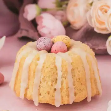 Mini bundt cake with white icing, topped with candy eggs, surrounded by pastel flowers on a pink surface.