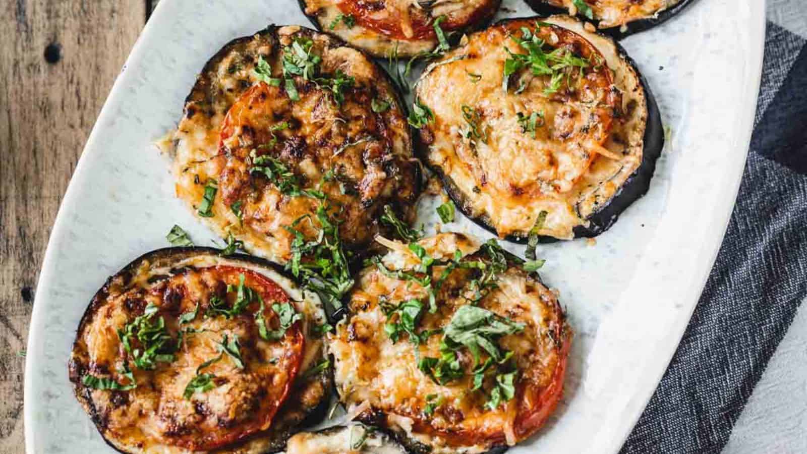 Slices of baked eggplant topped with tomato, melted cheese, and fresh herbs on a white plate.