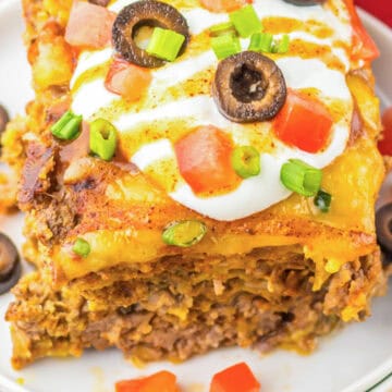 A slice of taco casserole, inspired by slow cooker recipes, is topped with sour cream, cheese, olives, tomatoes, and green onions on a white plate.