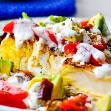 Grilled wedge of lettuce topped with diced tomatoes, avocado, and creamy dressing on a white plate.