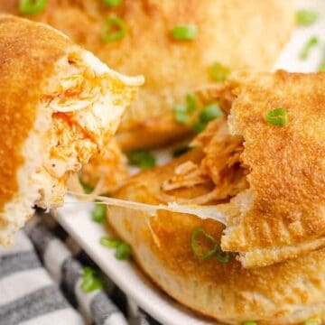 Hand holding a cheesy, shredded chicken-stuffed baked calzone with more calzones on a plate, garnished with green onions.
