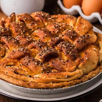 Golden-brown savory pie topped with seeds, with eggs and shredded cheese in the background on a wooden table.