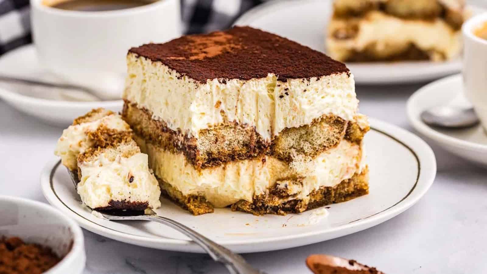 A slice of tiramisu on a white plate with a forkful taken out, surrounded by coffee cups.