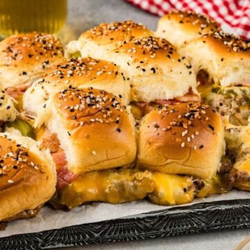 A tray of cheesy pulled beef sliders on sesame buns, perfect for Memorial Day recipes, with pickles and refreshing drinks in the background.