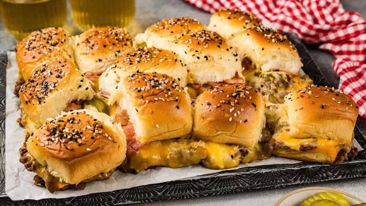 A tray of cheesy pulled beef sliders on sesame buns, perfect for Memorial Day recipes, with pickles and refreshing drinks in the background.