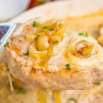 A spoon lifts a creamy pork chop topped with cooked onions and parsley from a baking dish.