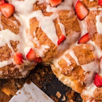 Close-up of glazed cinnamon rolls topped with fresh strawberry pieces in a skillet, one roll partially served.