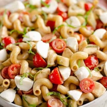 Bowl of pasta salad with cherry tomatoes, mozzarella, and fresh basil, garnished with lemon in the background.