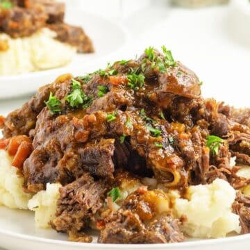 Shredded pot roast with gravy served over mashed potatoes, garnished with chopped parsley on a white plate.