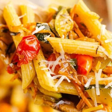 A close-up of penne pasta with vegetables, cherry tomatoes, and shredded cheese on a wooden spoon.