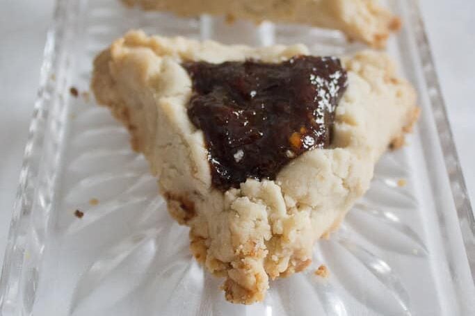 Two triangular pastries with a crumbly beige crust and dark prune filling, these vegan hamantaschen rest on a clear glass dish adorned with floral patterns. The elegant dish is set on a white, textured surface.
