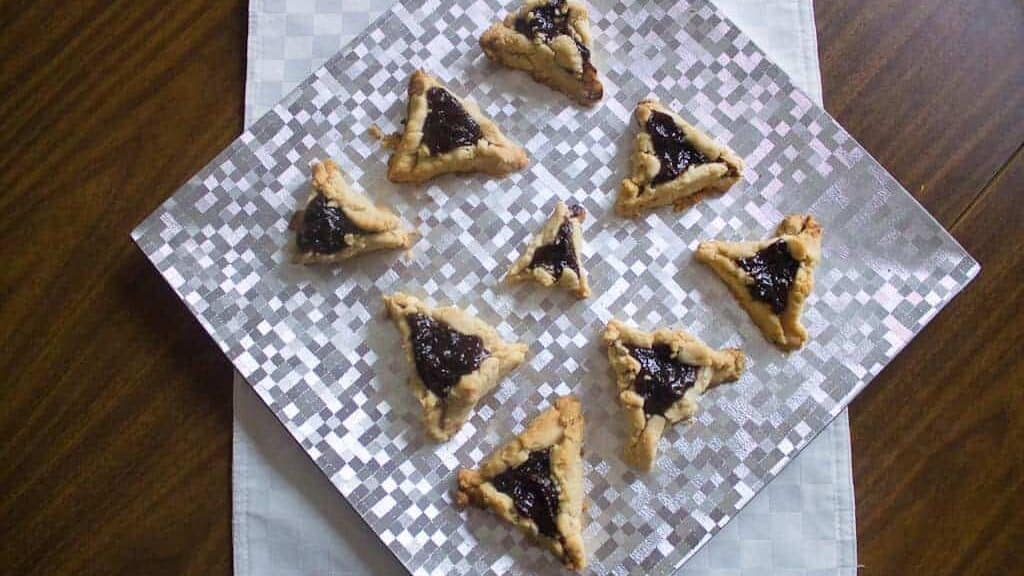 A square silver tray on a white cloth displays eight triangular prune hamantaschen with dark filling. The tray rests on a wooden surface.