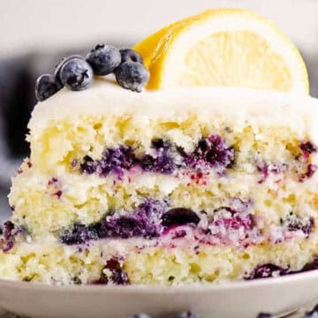 A slice of blueberry lemon cake with frosting, topped with blueberries and a lemon slice on a white plate.