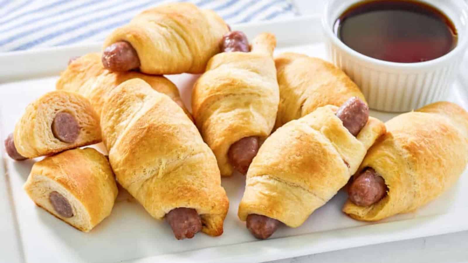 Golden brown crescent rolls wrapped around sausages on a plate, with a small bowl of dipping sauce&mdash;these retro appetizers are perfect for parties.