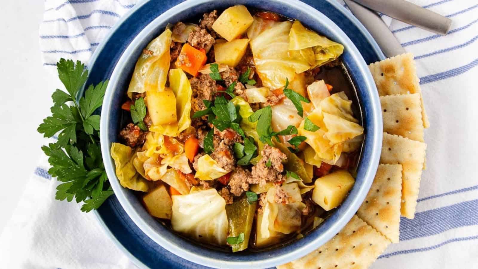 A bowl of cabbage soup with ground meat, potatoes, carrots, fresh herbs, and crackers on the side.