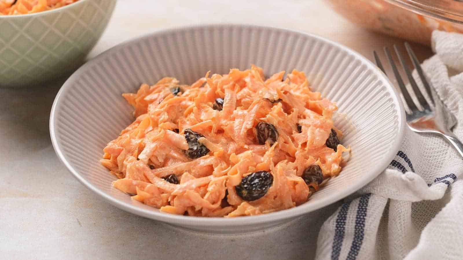 Shredded carrot salad with raisins and creamy dressing served in a white bowl on a light surface.
