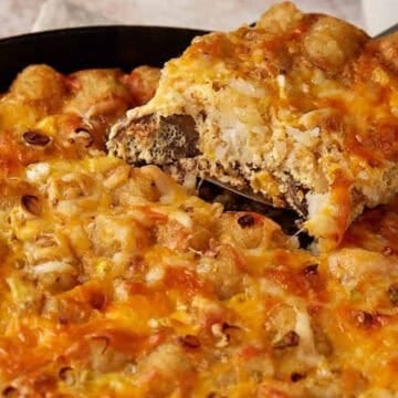 A cheesy tater tot casserole in a skillet, with a serving being lifted out.