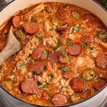 A pot of gumbo with sausage, shredded chicken, okra, and herbs, with a wooden spoon stirring it.