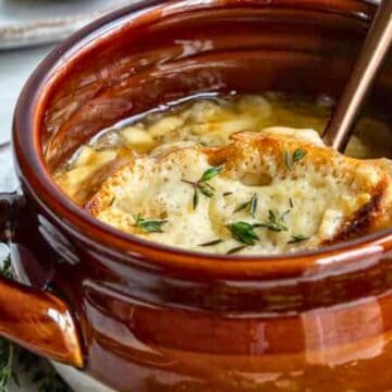Brown soup bowl of French onion soup topped with toasted bread and melted cheese, garnished with herbs.