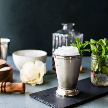 A silver julep cup with crushed ice and mint, surrounded by bar tools, a rose, and fresh mint jars.