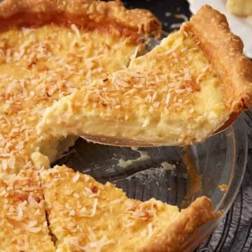 A slice of coconut cream pie—a classic among vintage desserts—is being lifted from a glass pie dish, topped with toasted coconut flakes.