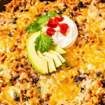 A skillet filled with cheesy taco casserole, topped with avocado slices, sour cream, cilantro, and diced tomatoes.
