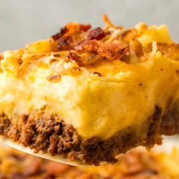 A close-up of a spatula holding a slice of shepherd's pie topped with cheese and crispy bacon.