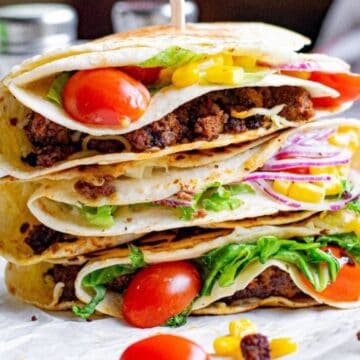 Three stacked folded tortillas filled with ground beef, lettuce, tomatoes, corn, and red onion on a white surface.