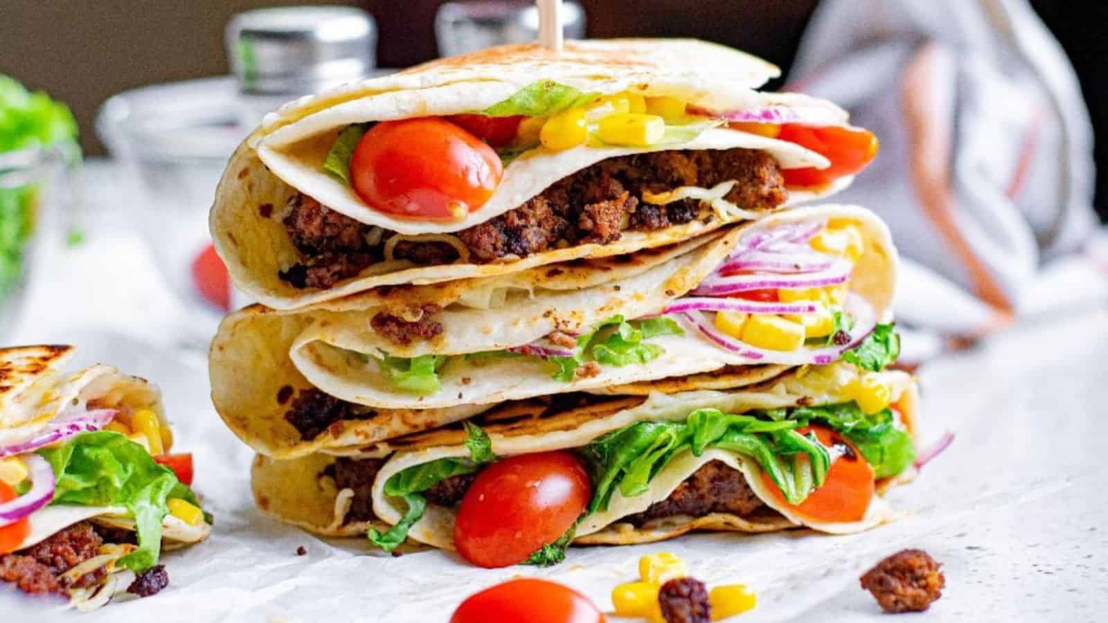 Three stacked folded tortillas filled with ground beef, lettuce, tomatoes, corn, and red onion on a white surface.