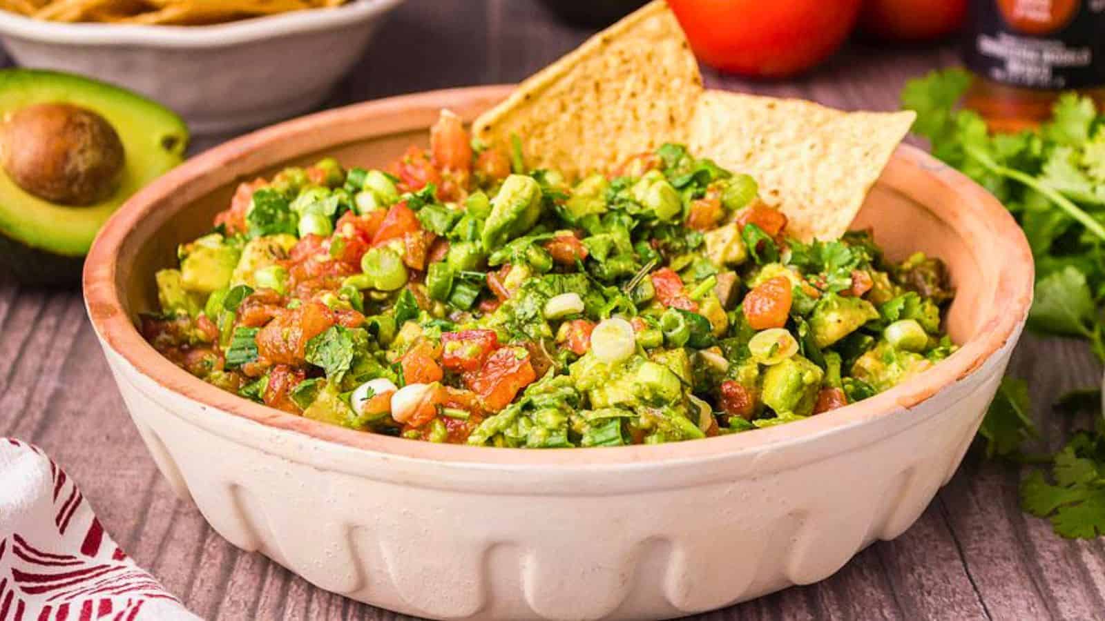 A bowl of guacamole with diced vegetables and two tortilla chips, placed on a wooden table.