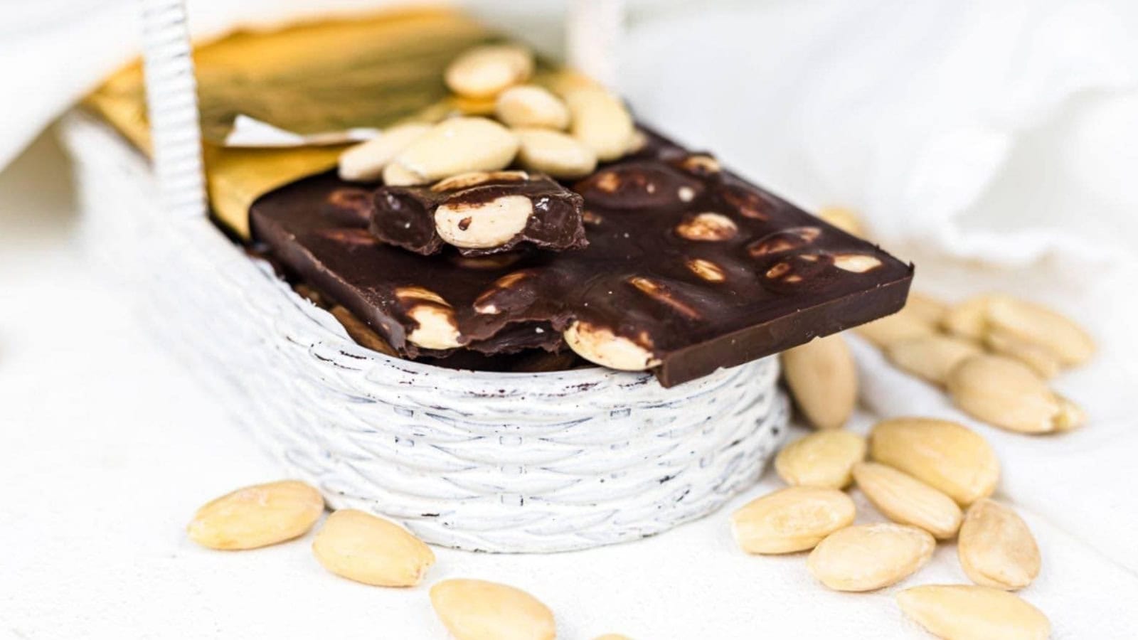 A bar of dark chocolate with nuts inside a small white woven basket, surrounded by scattered almonds on a white surface.