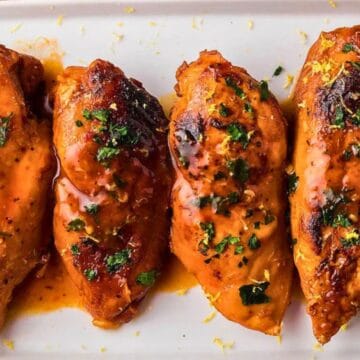 Four glazed chicken breasts garnished with parsley and lemon slices on a white rectangular plate.
